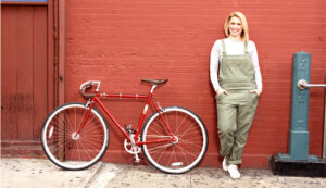 Dr. Alterman wears overalls, hands in pockets, leans against a red brick wall. Next to her, a red vintage road bike.