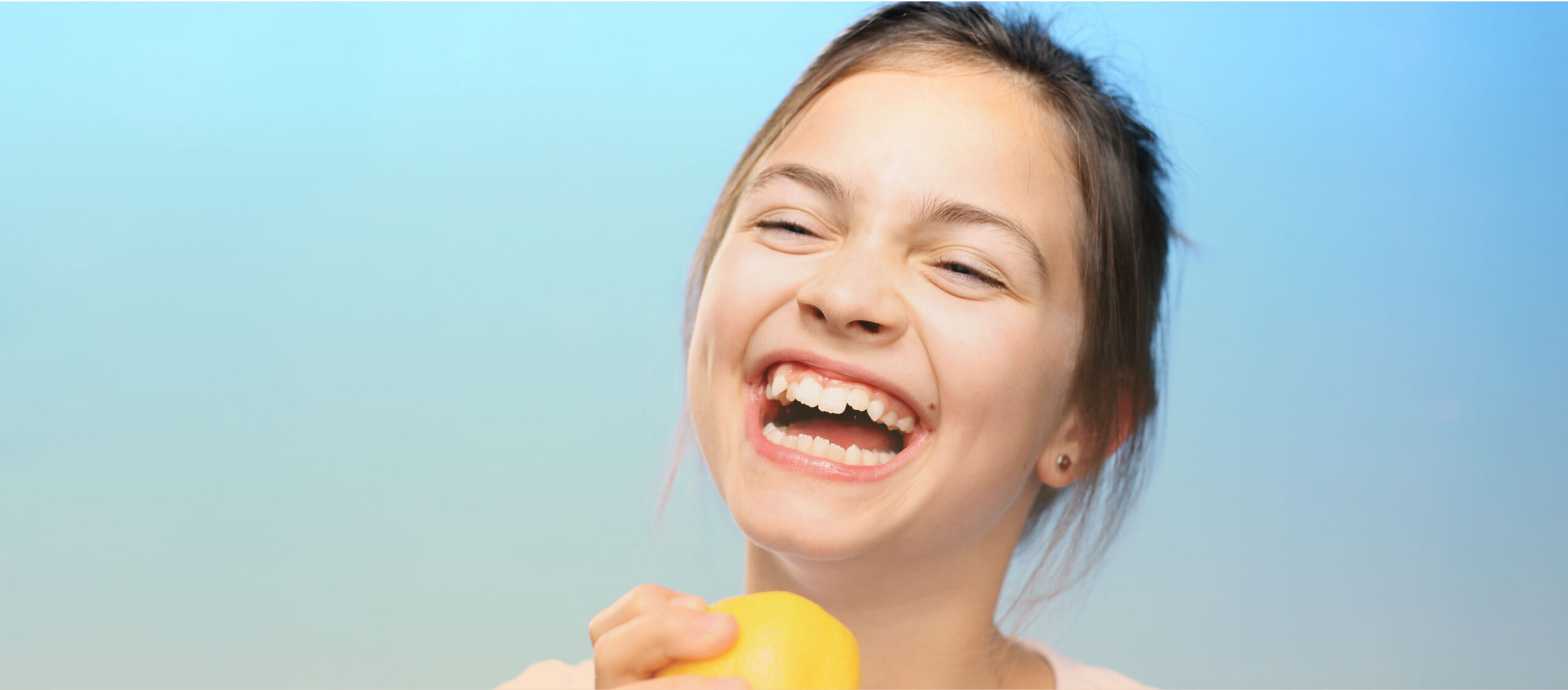 Tween girl with a piece of fruit in her hand laughs, tossing her head back.
